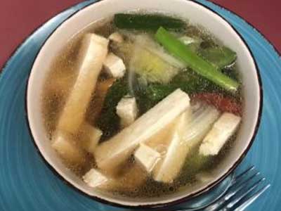 Top‑down view of vegetable tofu soup in clear broth with tofu strips, leafy greens and mixed vegetables, white bowl resting on blue plate, metal fork placed beside bowl, maroon table background.