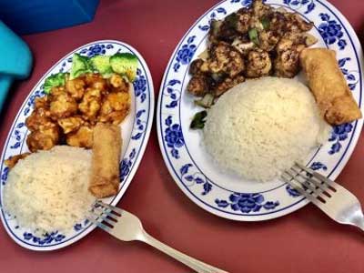 Two combination dinner plates on maroon table; left plate orange glazed chicken, broccoli, rice and egg roll; right plate black pepper chicken, steamed rice and egg roll, blue floral ceramic plates with metal forks.
