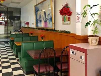 Interior dining room of Chinese restaurant, green vinyl booth seating, black‑and‑white checkered tile floor, wall artwork and festive decor, potted plant by host stand, glass entrance doors in background.