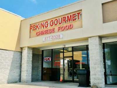 Storefront exterior of Peking Gourmet Chinese Food, red letter signage above glass entrance doors, stone support pillars, blue sky overhead, open sign displayed on front glass door.